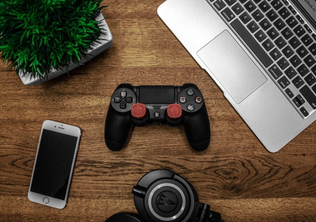 A black game controller on a wooden desk surrounded by a laptop, smartphone, headphones, and a small potted plant.